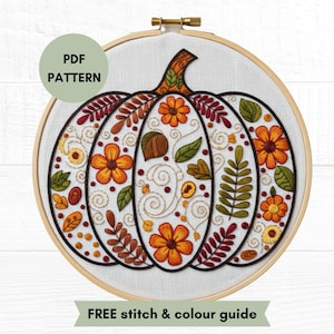 May include: A round wooden embroidery hoop containing white fabric with a hand-stitched pumpkin design. The pumpkin features orange flowers, green leaves, and brown stems with swirling gold thread accents. Text reads PDF PATTERN and FREE stitch & colour guide.