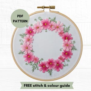 Könnte beinhalten: Ein runder Holzstickrahmen mit weißem Stoff, der einen handgestickten Kranz aus rosa und hellrosa Kosmeenblüten sowie grünen Blättern zeigt. Ein runder Aufkleber zeigt PDF PATTERN und ein unteres Banner trägt die Aufschrift FREE stitch & colour guide.