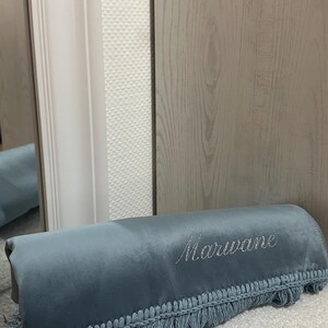 Op de afbeelding: Een opgevouwen, stoffig blauwe fluwelen deken met decoratieve franjes aan de onderkant. De naam Marwane is in cursief zilverkleurig garen op de voorzijde van de stof geborduurd, wat een persoonlijk detail aan dit item toevoegt.