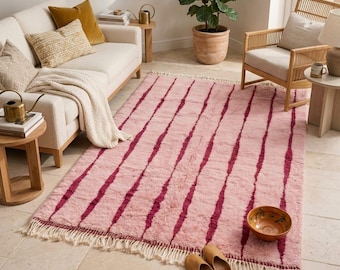 May include: A pink and burgundy striped rug with a fringed edge. The rug is placed in a living room setting, alongside a cream-colored sofa, wooden chair, and side tables. The room is decorated with pillows, a blanket, and a potted plant.