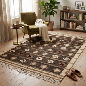 Può includere: Un tappeto rettangolare con un motivo geometrico nei toni del marrone, beige e rosso. Il tappeto ha un bordo con frange ed è posizionato su un pavimento in legno. La stanza comprende anche una poltrona verde, un tavolino e una libreria.