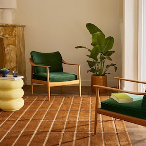 Op de afbeelding: Een rechthoekig vloerkleed met een rasterpatroon van bruine vierkanten gescheiden door dunne witte lijnen. Het kleed ligt op een houten vloer en dient als basis voor twee houten fauteuils met donkergroene fluwelen kussens en een gele bijzettafel.