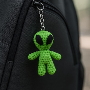 May include: A green crocheted alien keychain with black eyes and a silver chain attached to a black backpack zipper. The alien has a simple, rounded design.