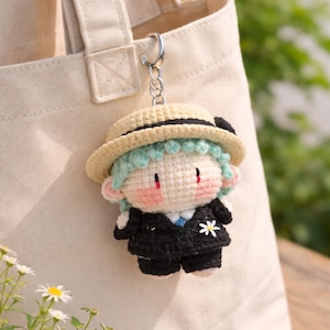 May include: A handmade crochet keychain featuring a doll-like figure with light green hair, a beige hat, and a black suit. The doll has rosy cheeks and a small white flower on the lapel. The keychain is attached to a silver ring.