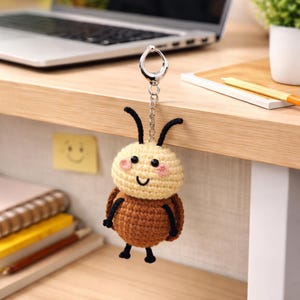 Puede incluir: Un llavero de ganchillo hecho a mano con forma de abeja. La abeja tiene una cabeza color crema con mejillas rosadas, ojos negros y una boca sonriente. El cuerpo es marrón y tiene antenas y patas negras. El llavero está sujeto a un cierre de metal plateado.