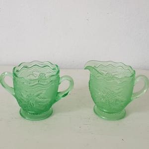 May include: A vintage green glass sugar bowl and creamer set with a floral design. The set has two handles and a scalloped edge.