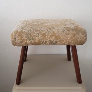 May include: A vintage footstool with a rectangular, cushioned top featuring a floral pattern in shades of gold and cream. The stool has four dark brown, tapered wooden legs. The footstool is a decorative piece.
