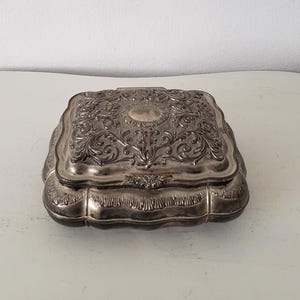 May include: A silver trinket box with an ornate floral design and a raised circular center. The box has a hinged lid and a slightly rounded shape.