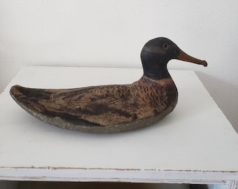 May include: A vintage wooden duck decoy with a black head and a brown and black painted body. The decoy is lying on a white surface.