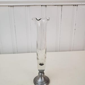 May include: A clear glass bud vase with a ruffled top and etched leaf design. The vase is mounted on a silver-colored metal base. The vase is approximately 9 inches tall.