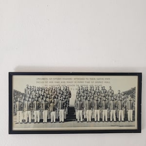 May include: A framed black and white photograph featuring a group of uniformed soldiers. The image includes text at the top, and the soldiers are standing in rows. The photograph is mounted on a white wall.