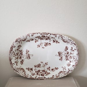 May include: An oval serving platter with a white background and a brown floral pattern. The rim features a decorative border. The platter is made of ceramic and is suitable for serving food.