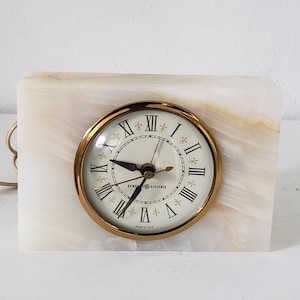 May include: A white marble clock with gold accents. The clock face has Roman numerals and a gold-colored General Electric logo. The clock is made in the USA.