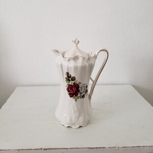 May include: White ceramic creamer with a gold trim and a floral design featuring red and white roses.