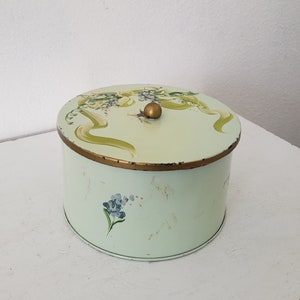 May include: A vintage, round, green metal tin with a gold lid and floral design. The lid has a gold knob and the tin has a floral design with green leaves and white flowers. The tin has a faded, distressed look.