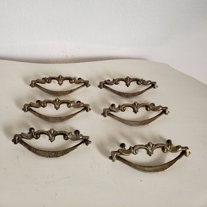 May include: Eight antique brass drawer pulls with ornate designs. The pulls have a curved shape and are approximately 3 inches long.