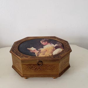 May include: A vintage, gold-toned, octagonal jewelry box. The lid features an oval painting of a woman reading. The box has ornate floral and scrollwork details, a small metal clasp, and rests on four small feet. A decorative storage item.