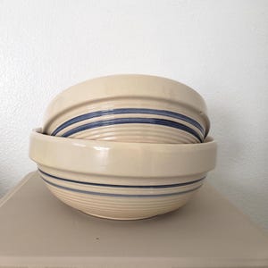 May include: Two cream-colored ceramic bowls stacked on top of each other. Each bowl has a wide, dark blue stripe design around the middle. The bowls have a glossy finish and are sitting on a light beige surface.