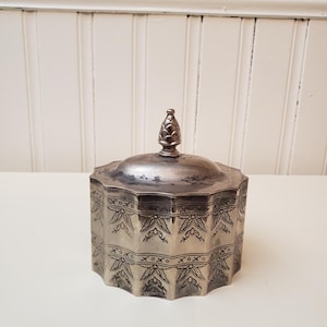 May include: A silver-toned, octagonal box with a lid and a decorative finial. The box features an etched design around the sides. The lid has a small, cone-shaped handle. The box is likely used for storing small items.