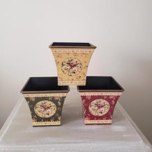 May include: Three square planters with a flared top edge and a black interior. The top planter is cream, the bottom left is dark green, and the bottom right is deep red. Each features a central circular floral medallion and floral borders.