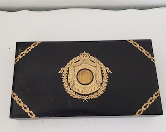 May include: A black rectangular box with gold trim and a gold design of a crown and wreath. The design includes a small circular image of a person's face.