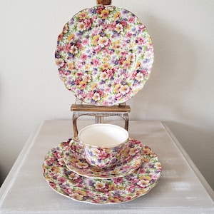 May include: A vintage floral teacup, saucer, and plate set. The set features a vibrant pattern of pink, yellow, and purple flowers on a white background, with gold trim. A larger plate is displayed behind the set on a small decorative stand.