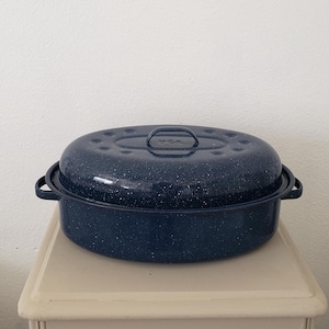 May include: A navy blue speckled enamel roasting pan with a matching lid. The oval-shaped pan has handles on each side and a handle on the lid. The pan is sitting on a cream-colored surface.