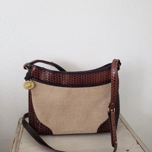 May include: A brown and tan shoulder bag with a woven pattern and a long strap. The bag features a tan center panel and dark brown woven leather accents. A gold-colored emblem hangs from the top left corner.