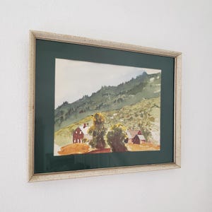 May include: Framed watercolor painting of a rural landscape. The artwork depicts a hillside with green trees and a few red-roofed buildings. The painting is set against a dark green mat and framed in a light-colored, textured frame.