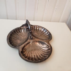 May include: A silver-toned, three-section serving dish with a handle. Each section has a shell-like design. The handle is ornate and connects to the dish's edge. The dish is on a white surface.