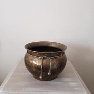 May include: A round brass planter with a polished finish and a dark patina. The upper rim features a series of small indentations, and a decorative twisted brass rope detail encircles the upper body, ending in two small hanging ball accents.