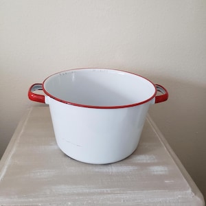 May include: A white enamel cooking pot with a bright red rim and two matching red side handles. The pot features a smooth, glossy finish with a wide, circular opening and a deep basin, showing minor signs of wear along the upper red edge.