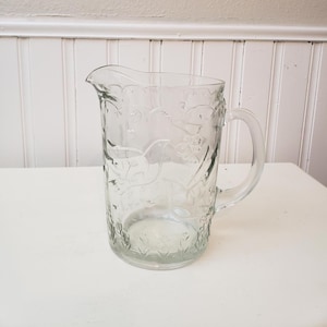 May include: A clear glass pitcher featuring an embossed design of birds and floral patterns. The vessel has a curved handle, a wide pouring spout, and a scalloped rim. The surface texture is smooth with raised decorative elements throughout.