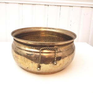 May include: A vintage brass planter with a rounded shape and a decorative rope detail around the middle. The planter has a shiny, aged finish and a rope handle. The interior is visible, suggesting it's ready for plants or decorative items.