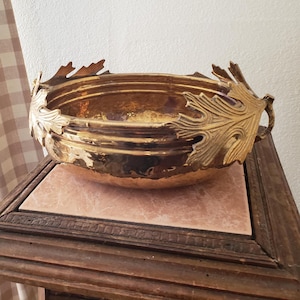 May include: A decorative brass bowl with a hammered texture and a leaf-shaped rim. The bowl is round and sits on a wooden stand with a pink tile top. The leaves are a bright gold color.