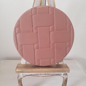 May include: A round, dusty rose-colored decorative piece with a woven pattern, displayed on a small, ornate wooden easel. The easel has a gold-painted top and distressed white and gold legs, creating a vintage aesthetic.