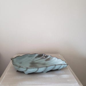 May include: A leaf-shaped ceramic dish featuring a light blue glaze with dark gray accents along the edges and center. The dish has a scalloped rim and small feet on the underside, providing a shallow basin for holding small items or trinkets.