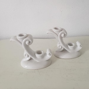 May include: Two white ceramic candle holders with a decorative, swirling design. Each holder has a single candle cup.