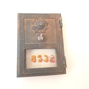 May include: Antique bronze-colored mailbox door with a decorative star-shaped knob and a small window displaying the number "8332" in red and yellow. The door has a rectangular shape with a patterned border.