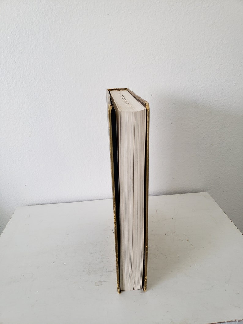 May include: A gold-colored book with a white spine and a black cover. The book is standing upright on a white surface.