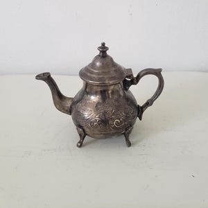 May include: A silver teapot with an ornate design and three legs. The teapot has a lid with a knob on top.