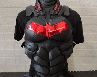 May include: A black tactical chest armor piece featuring a textured carbon fiber pattern and a prominent metallic red bat-shaped emblem on the chest. The armor includes shoulder guards and collar detailing, with the text SapnahCosplayStore visible.