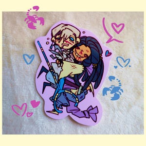 May include: A die-cut sticker featuring a stylised illustration of two characters embracing. One character has blonde hair and the other has dark hair. The design includes pink and blue scorpion icons, heart shapes, and a light purple background.