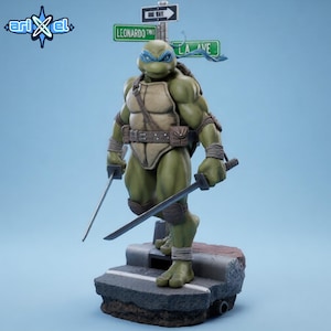 May include: A detailed figurine of Leonardo from Teenage Mutant Ninja Turtles, standing on a stone base. The figure is green with a blue mask, holding two katanas. Street signs read "Leonardo" and "LA AVE". The Arikel logo is in the upper left corner.