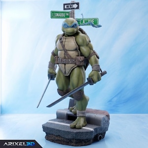 May include: A green Teenage Mutant Ninja Turtle figurine wearing a blue mask and brown gear, holding two katana swords. The figure stands on a grey street-themed base with a street sign post behind it labelled Leonardo and L.A. Ave.