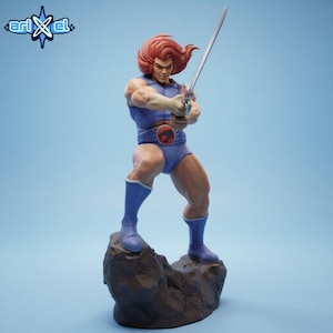 May include: A detailed figurine of a muscular character from the Thundercats series, holding a sword. The figure is clad in a blue outfit with a red and black belt buckle. The character has red hair and is standing on a rocky base against a light blue backdrop.
