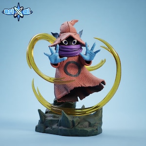 Puede incluir: Figura detallada de un personaje con túnica roja, bufanda morada y guantes azules. La figura tiene un sombrero marrón y está rodeada por un efecto de energía amarilla. La base es un terreno rocoso, y el logo "ari x el" está en la esquina superior izquierda.