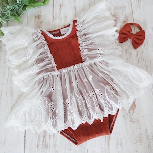 May include: A rust-colored baby romper with white lace detailing and a matching bow headband. The romper features a textured bodice and a delicate, embroidered lace skirt and sleeves. The headband has a bow.