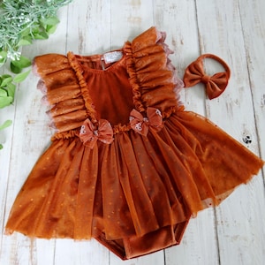 May include: Rust-colored baby romper with a matching headband. The romper features ruffled sleeves, delicate lace trim, and a tulle skirt with small polka dots. Two small bows adorn the bodice. The headband has a matching bow.
