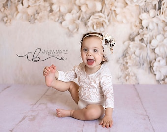 May include: A baby girl wearing a white lace bodysuit and a brown headband with a feather accent sits on a light purple floor in front of a white floral backdrop. She is smiling and looking at the camera.